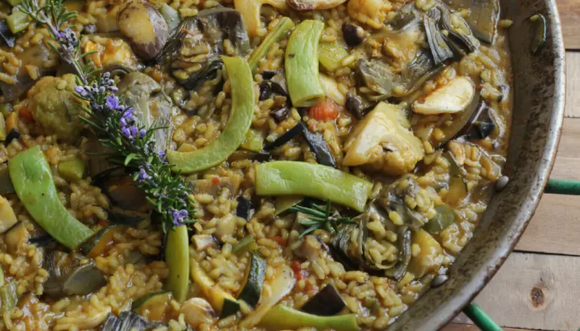 Paella de verduras vibrante, con alcachofas, calabacín, pimientos y romero fresco. Un festín para la vista y el paladar.