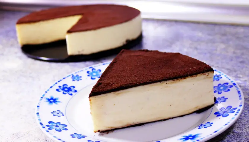 Un delicioso pastel de queso cubierto de cacao, perfecto para postres fáciles. Una rebanada servida en un plato con flores azules.