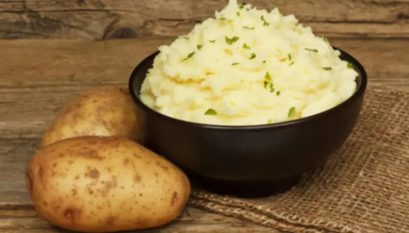Un cremoso puré de patatas Thermomix, adornado con perejil fresco, junto a patatas crudas sobre una mesa de madera.
