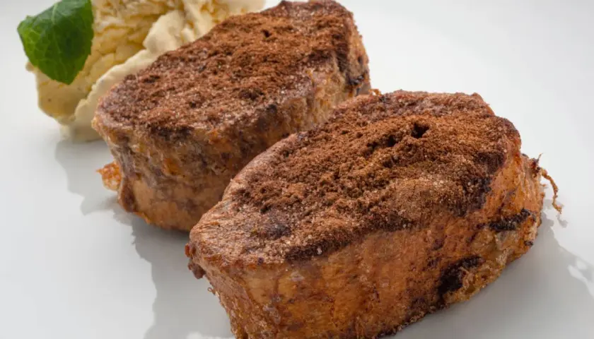 Dos torrijas de chocolate espolvoreadas con cacao y azúcar, acompañadas de helado de vainilla y una hoja de menta.