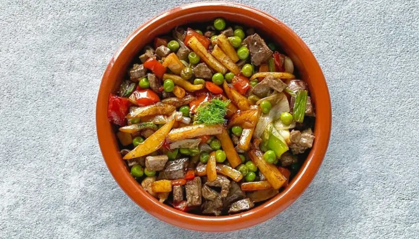 Un sabroso frito mallorquín con trozos de carne, patatas, guisantes y pimientos en una cazuela de barro.