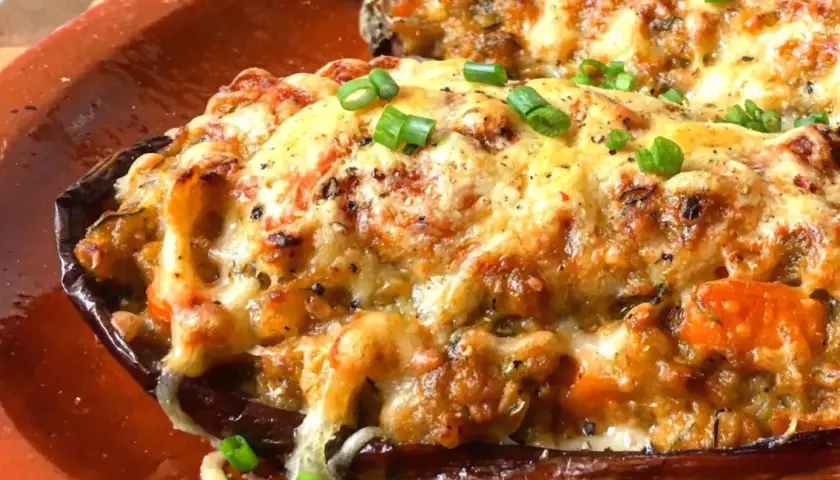 Berenjenas rellenas gratinadas con queso derretido y cebolleta picada, servidas en una cazuela de barro.