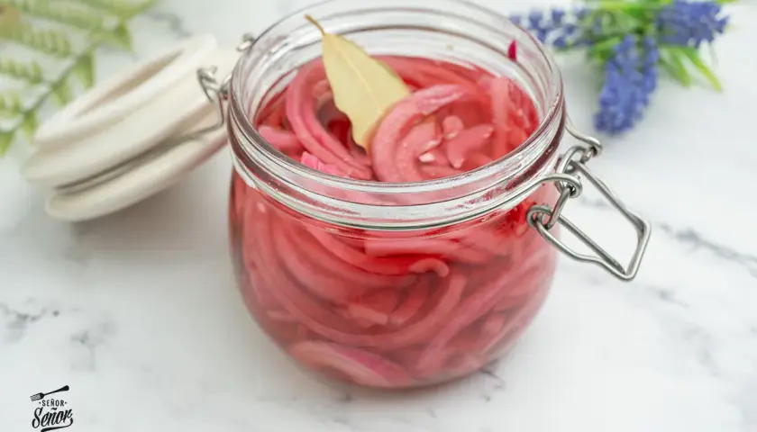Frasco de vidrio lleno de cebolla encurtida, con una hoja de laurel flotando. Un toque de color y sabor para tus platillos.