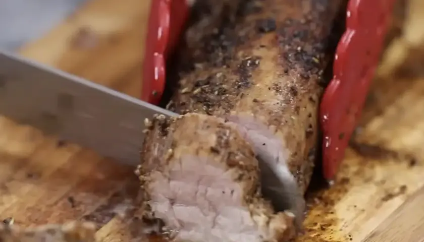 Un jugoso solomillo de cerdo al horno, recién cortado en una tabla de madera. Las pinzas rojas sujetan la carne mientras un cuchillo la rebana.