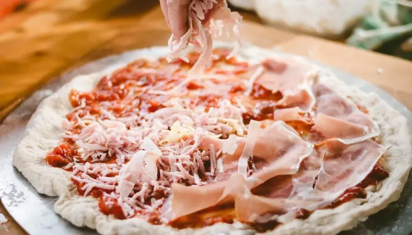 Manos esparciendo queso rallado sobre una pizza casera con jamón y salsa de tomate.