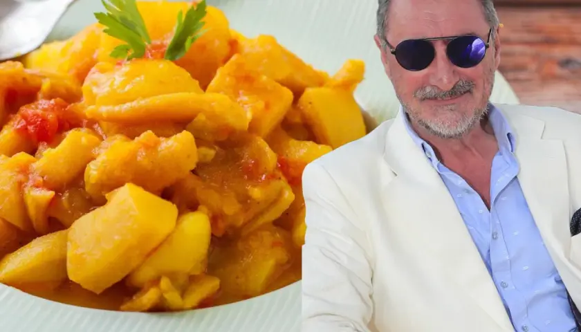 Un hombre con gafas de sol y traje blanco sonríe junto a un plato de papas con choco, un guiso amarillo con trozos de tomate y perejil.