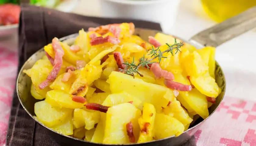 Patatas al microondas con trocitos de bacon crujiente y romero fresco, servidas en una sartén metálica.