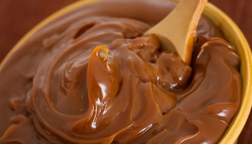 Cuchara de madera removiendo un tazón lleno de dulce de leche cremoso, perfecto para tu dulce de leche receta.