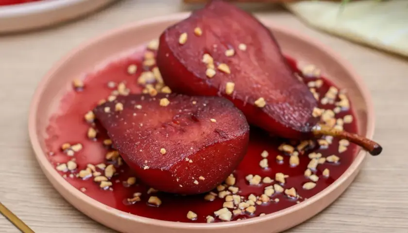 Peras al vino, tiernas y brillantes, servidas en un plato rosa con trocitos de nuez y un sirope rojo intenso.