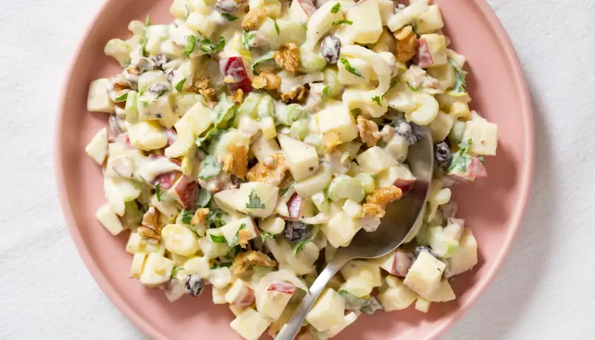 Ensalada Waldorf cremosa con trozos de manzana, apio, nueces y pasas, servida en un plato rosa.