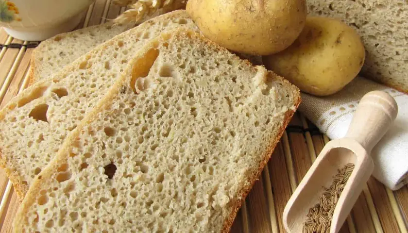 Rebanadas de pan de patata rústico, con patatas enteras, espigas de trigo y comino en una cuchara de madera.