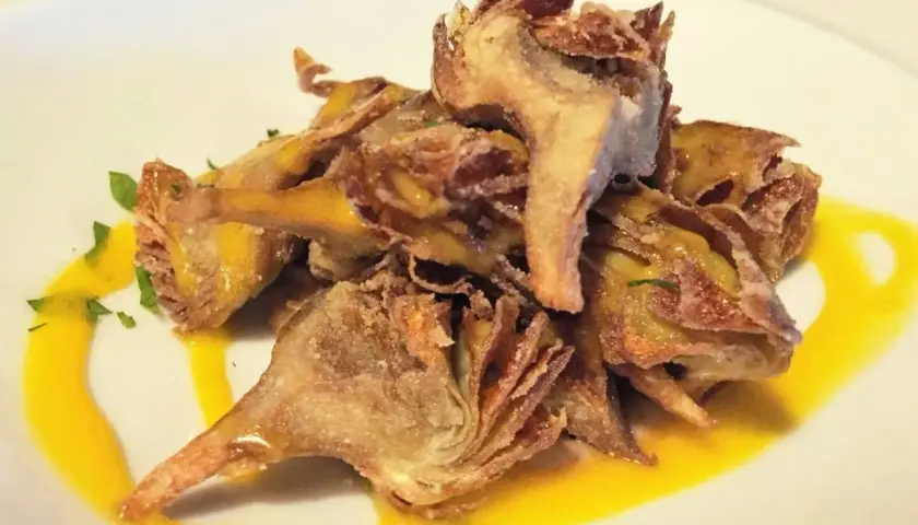 Montón de alcachofas fritas crujientes, doradas y sazonadas, servidas sobre un plato blanco con un toque de salsa amarilla y perejil picado.