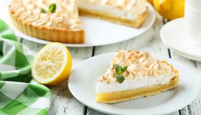 Delicioso pastel de limón con merengue dorado, decorado con menta fresca. Un trozo servido en plato blanco, junto a limones y una taza de té.
