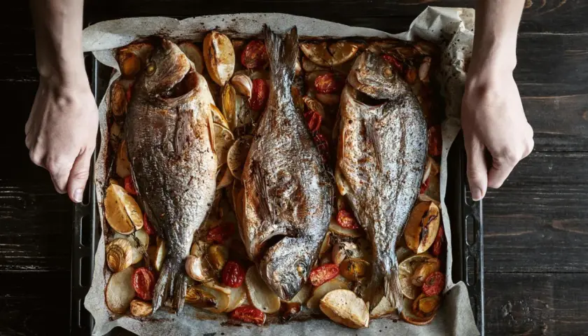 Tres pescados dorados al horno, acompañados de rodajas de limón, tomates cherry y ajos, listos para servir.
