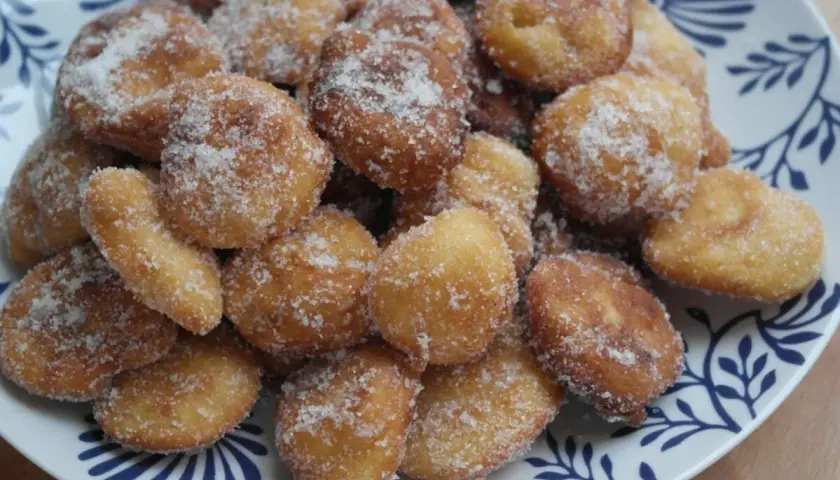 Montón de buñuelos de manzana dorados y espolvoreados con azúcar, servidos en un plato blanco con diseño azul.