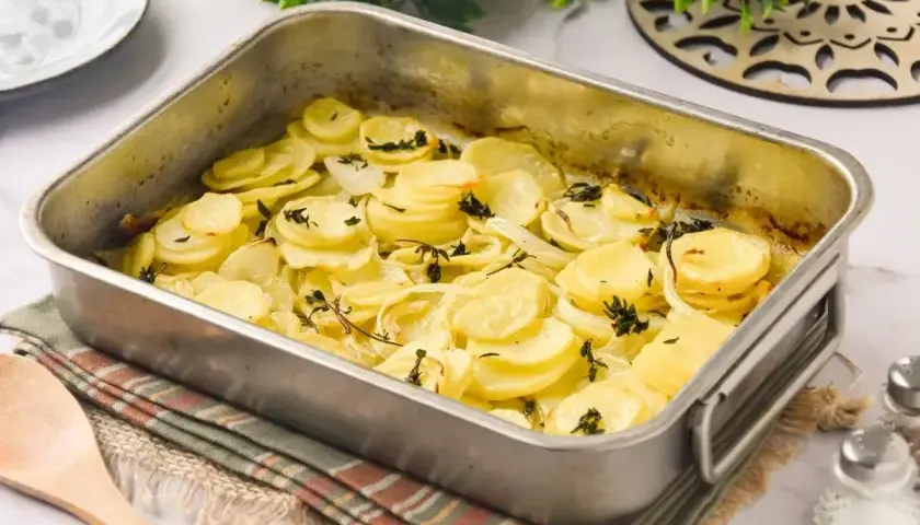Patatas a lo pobre recién horneadas en una bandeja metálica, con rodajas finas de patata y cebolla doradas y hierbas aromáticas.
