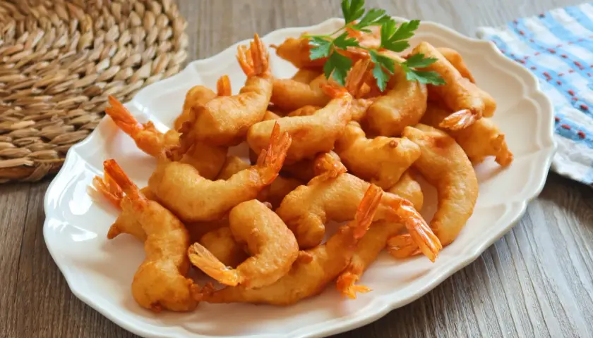 Un plato blanco lleno de gambas a la gabardina doradas y crujientes, decorado con perejil fresco.