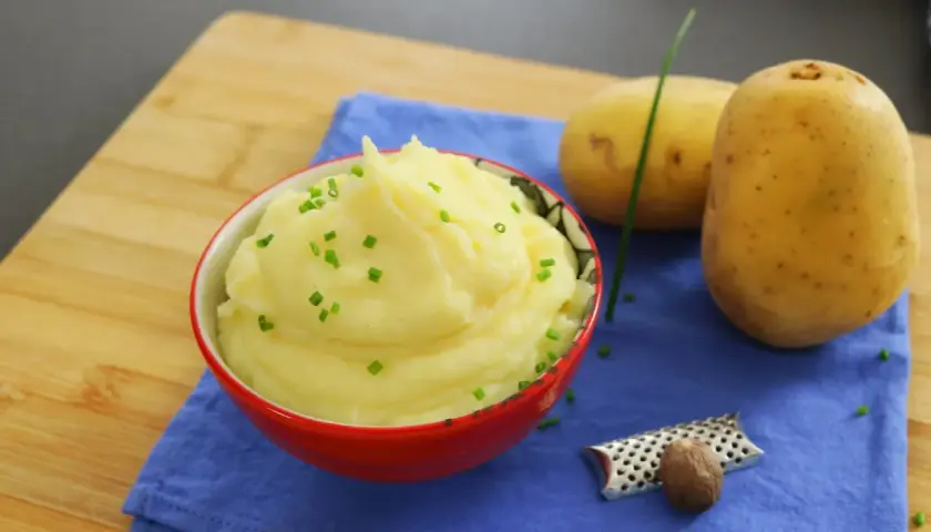 Un cremoso puré de patatas, adornado con cebollino fresco, junto a patatas crudas y una nuez moscada.