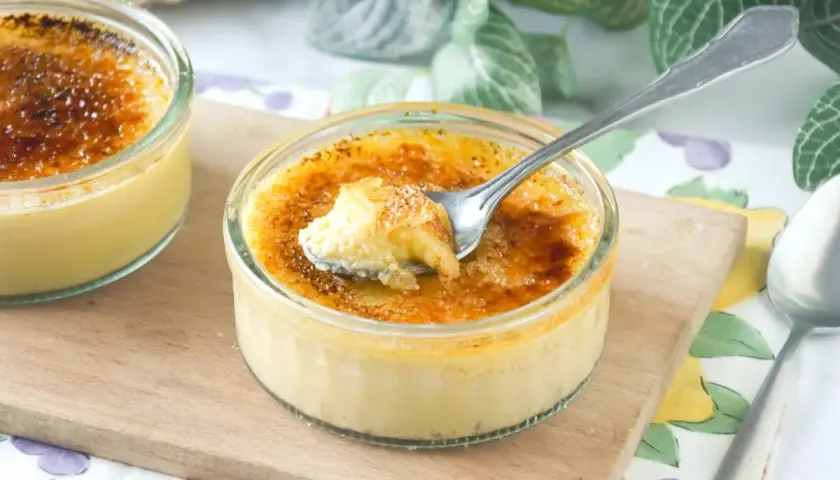 Dos cremosos postres de creme brulee en vasitos de cristal, uno con una cucharada sacada, sobre una tabla de madera.