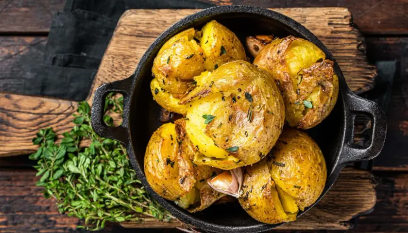 Patatas al horno doradas y crujientes, con hierbas y ajo, servidas en una sartén de hierro fundido sobre una tabla de madera.