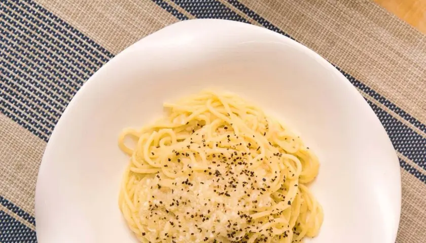 Un plato de cacio e pepe, pasta cremosa con queso y pimienta negra, servido en un cuenco blanco sobre un mantel a rayas.