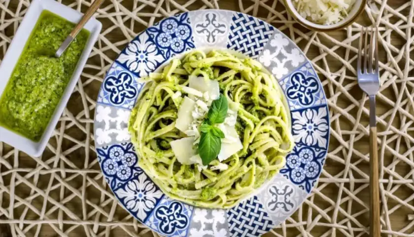 Un plato de pasta al pesto, decorado con queso parmesano y albahaca fresca, junto a una pequeña porción de salsa pesto.