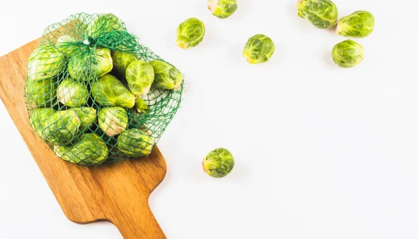 Una bolsa de malla verde llena de coles de Bruselas frescas descansa sobre una tabla de cortar de madera. Algunas coles de Bruselas están esparcidas alrededor.
