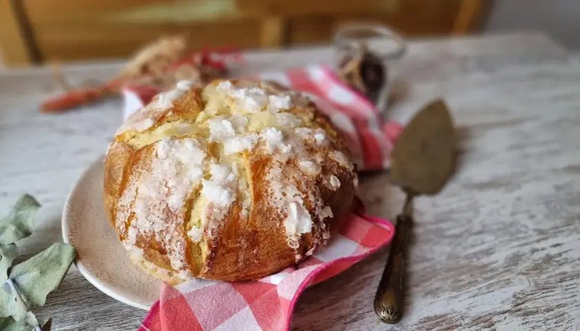 Un pan quemado dorado y crujiente, espolvoreado con azúcar, descansa sobre un plato. Una servilleta a cuadros rojos y blancos y un utensilio para pasteles completan la escena.
