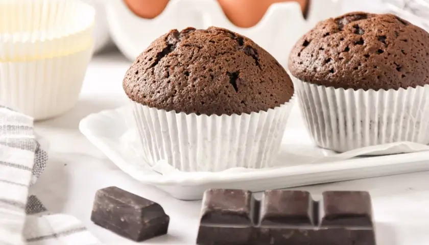 Dos magdalenas de chocolate recién horneadas, listas para disfrutar. A su lado, trozos de chocolate y moldes para hornear.