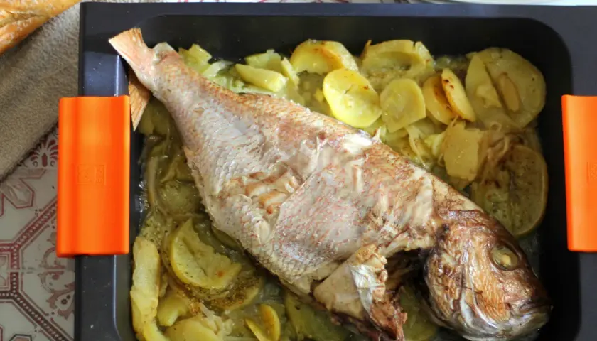 Pargo al horno con patatas y cebolla en una bandeja negra. Un pan rústico descansa a un lado.