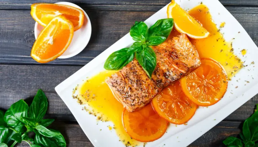 Delicioso salmón a la naranja, servido con rodajas de naranja y hojas de albahaca fresca.
