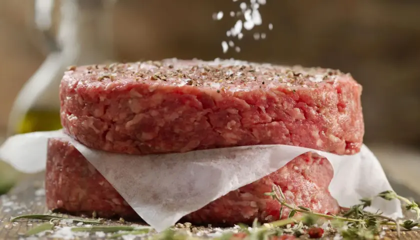Dos hamburguesas de carne picada apiladas, sazonadas con sal y pimienta.