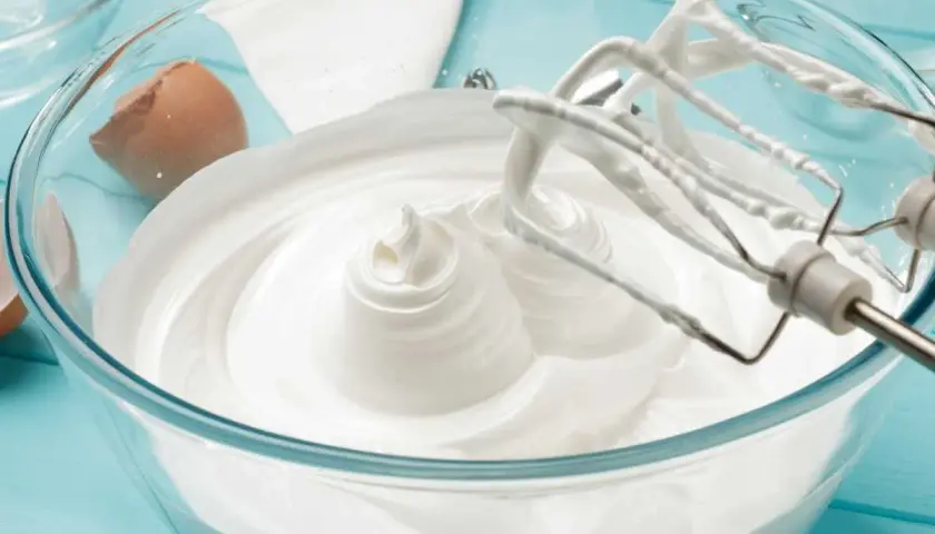 Batidora mezclando merengue italiano en un bol de cristal. Cáscara de huevo al lado.