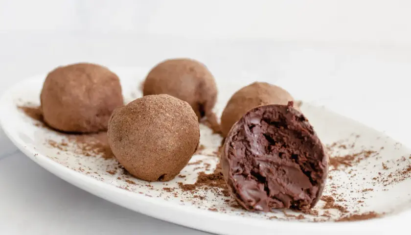 Cinco trufas de chocolate espolvoreadas con cacao en polvo sobre un plato blanco. Una trufa está partida, revelando su interior cremoso.