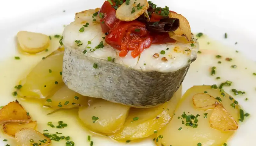 Deliciosa merluza a la plancha sobre patatas, con pimientos asados, ajo crujiente y perejil picado.