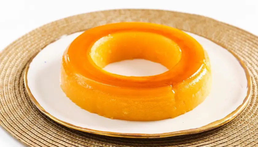 Un delicioso tocino de cielo, con su característico color dorado y forma de anillo, reposa sobre un plato blanco con borde dorado.