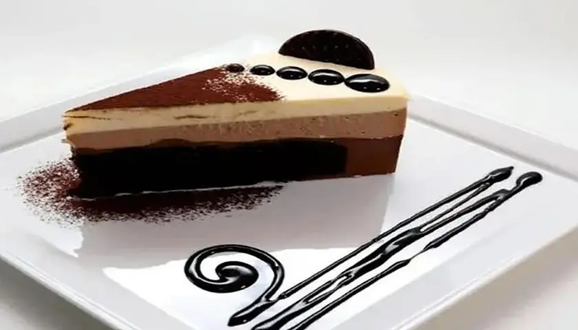 Una rebanada de tarta de tres chocolates, con capas de chocolate blanco, con leche y negro, decorada con cacao en polvo y un disco de chocolate.