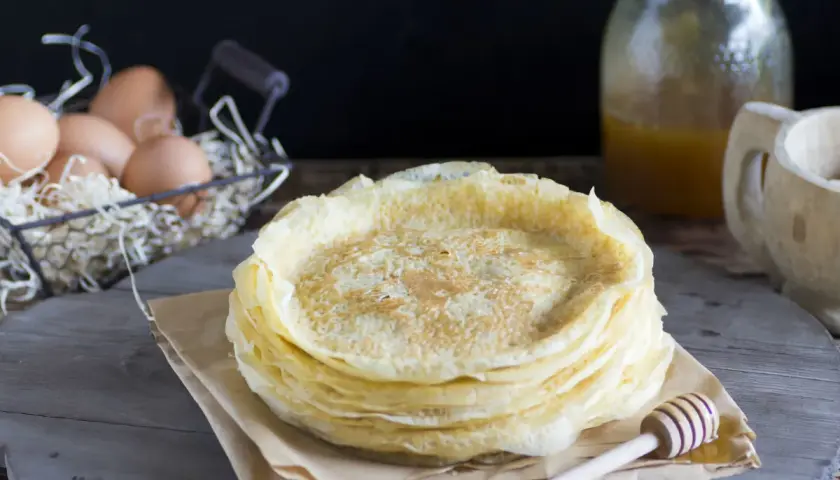 Pila de filloas gallegas recién hechas, con miel en un tarro y huevos en una cesta al fondo.