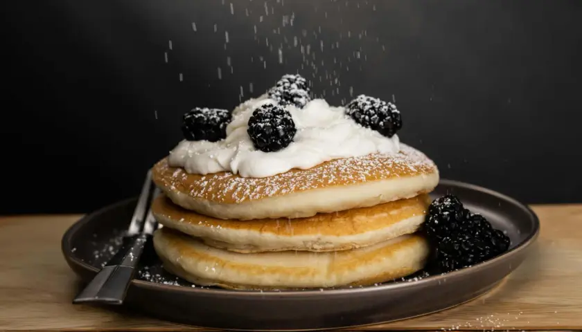 Una pila de tortitas americanas cubiertas con crema batida y moras, espolvoreadas con azúcar glas.