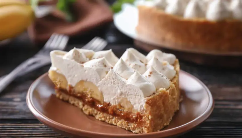 Rebanada de tarta banoffee con plátano, dulce de leche y crema batida, espolvoreada con canela.