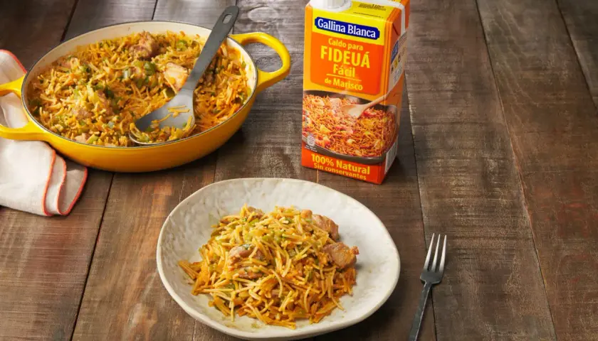 Fideos a la cazuela servidos en un plato y una paellera amarilla, junto a un caldo para fideuá.