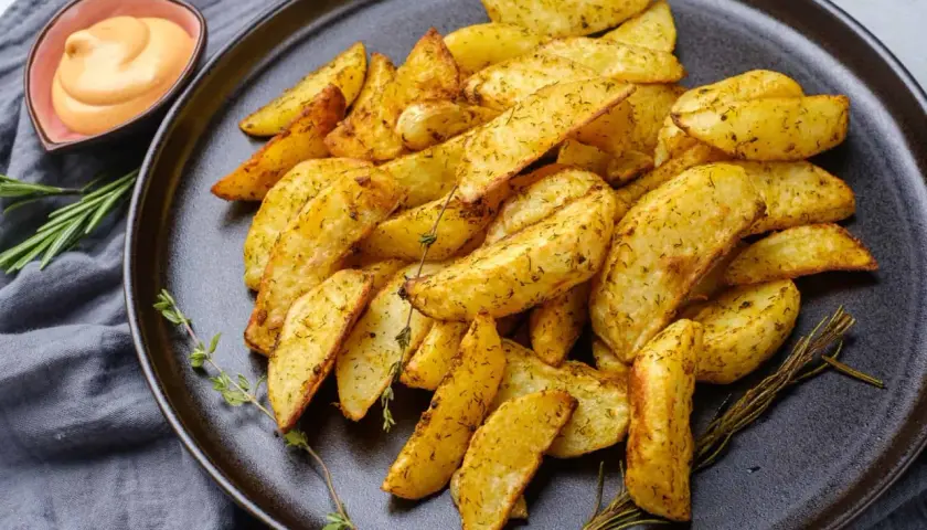 Patatas fritas al horno doradas y crujientes, sazonadas con hierbas, servidas en un plato oscuro con una salsa cremosa y romero fresco.