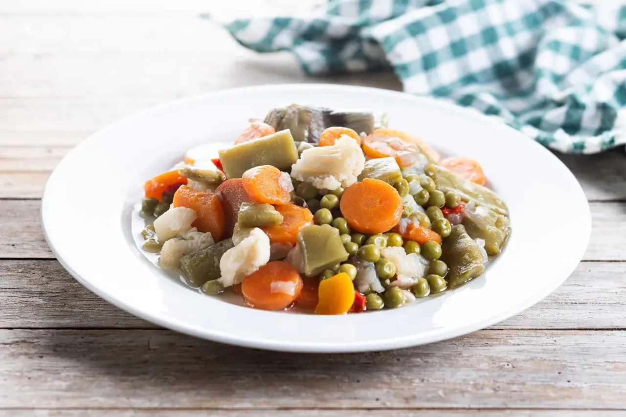 Un plato de menestra de verduras con zanahorias, guisantes, coliflor y judías verdes, servido en una vajilla blanca sobre una mesa de madera.