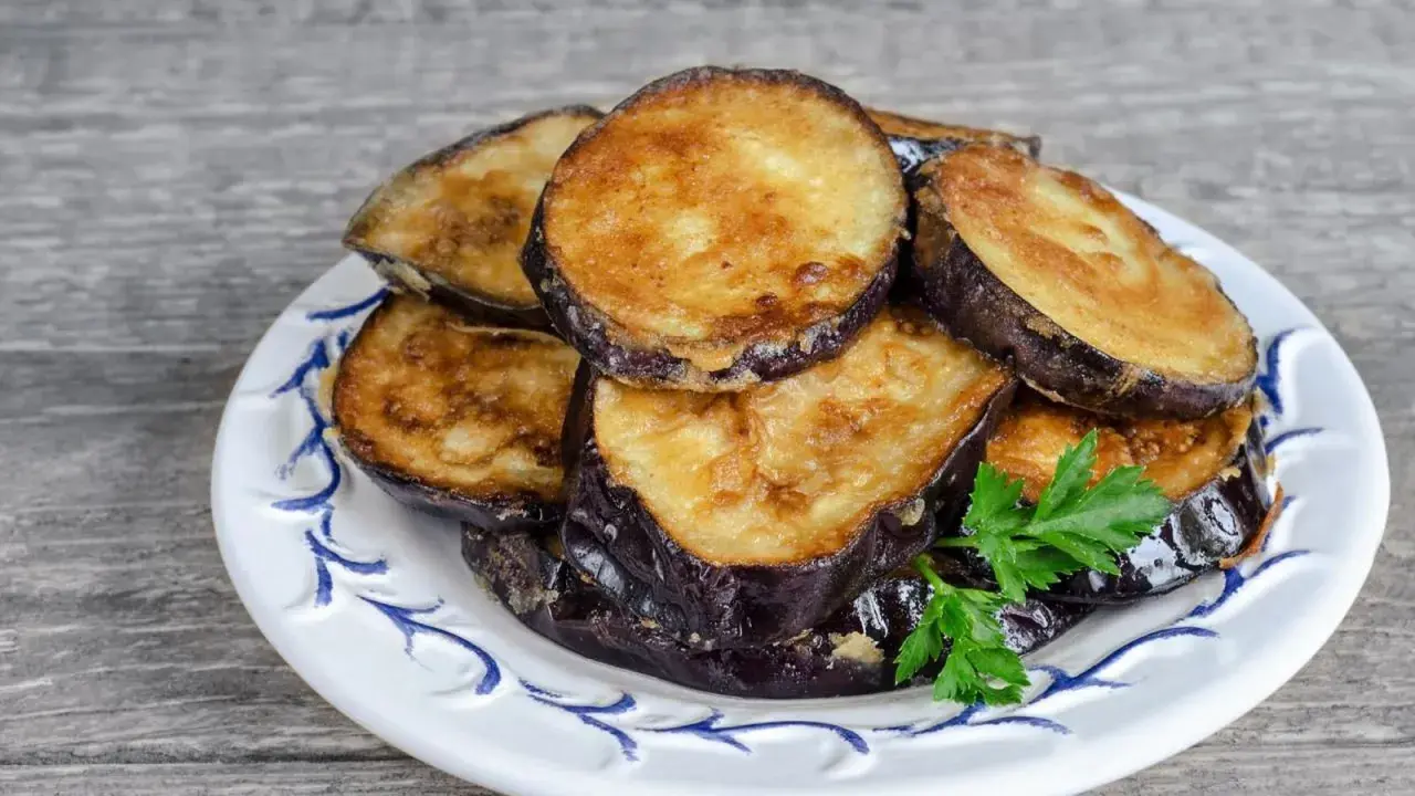 Rodajas doradas de berenjenas fritas apiladas en un plato blanco con decoración azul, adornadas con perejil fresco.