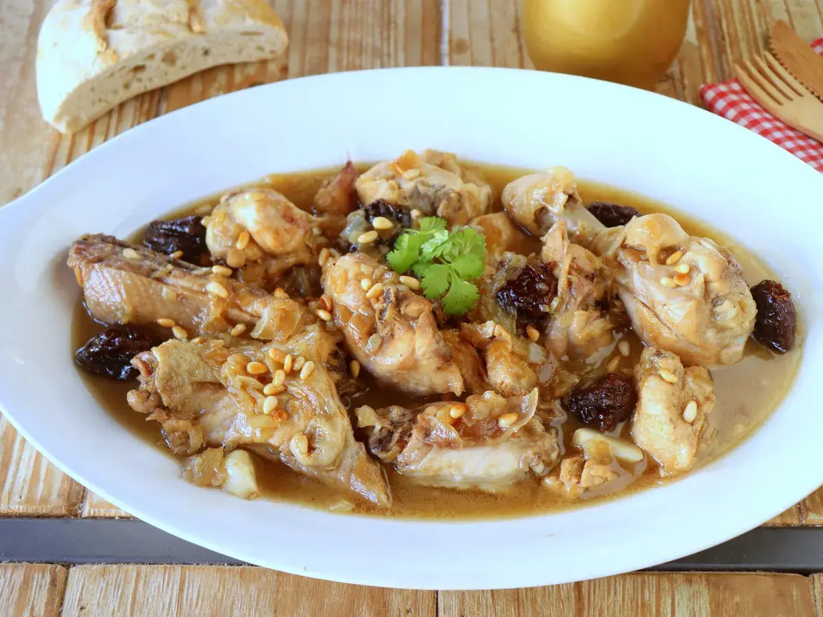 Delicioso pollo guisado con ciruelas pasas y piñones, servido en un plato blanco con pan rústico al fondo.