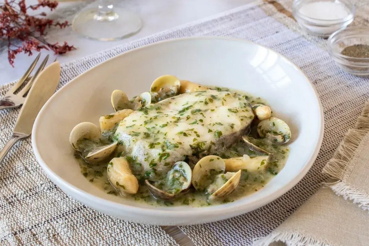 Delicioso pescado en salsa verde con almejas, servido en un plato blanco sobre un mantel de lino.