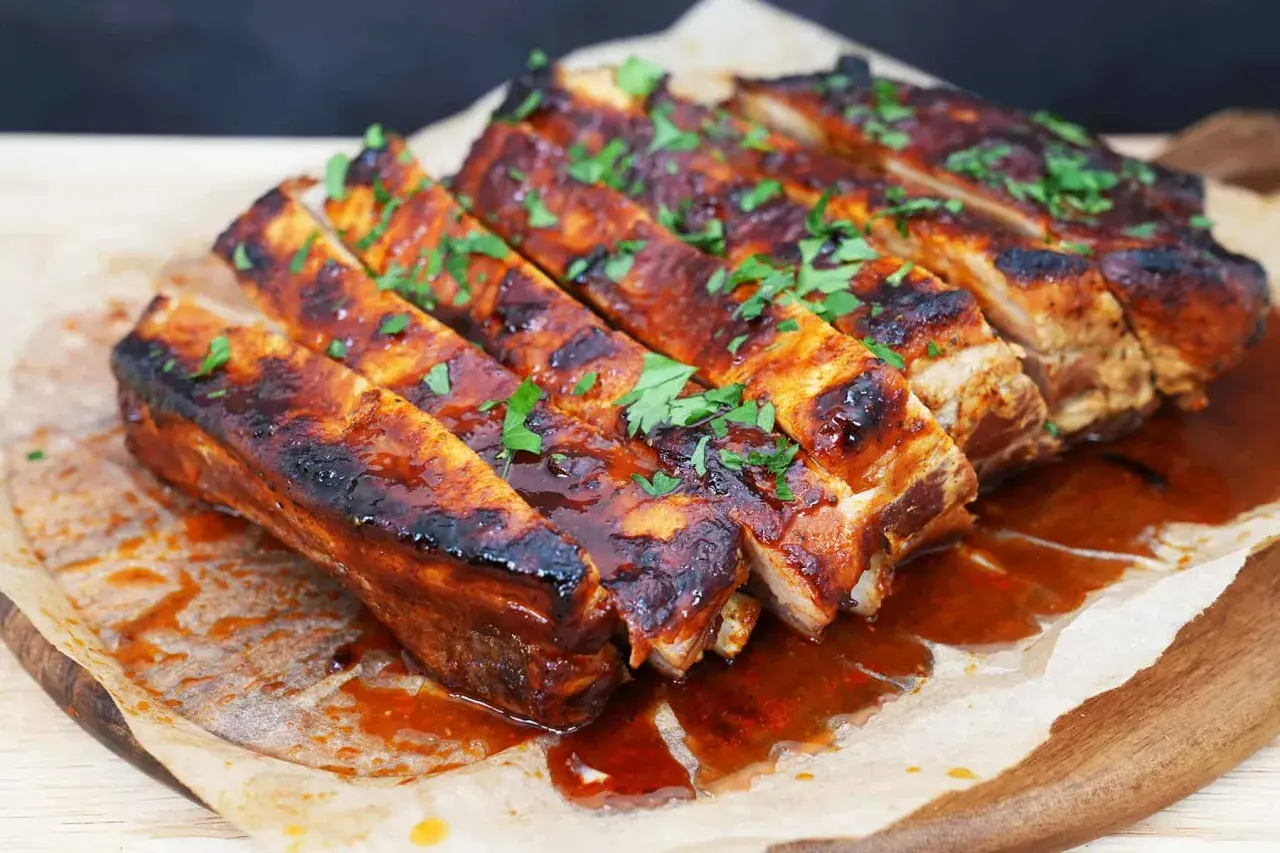 Costillas a la barbacoa jugosas y glaseadas, cubiertas con perejil fresco picado, servidas sobre papel de hornear en una tabla de madera.