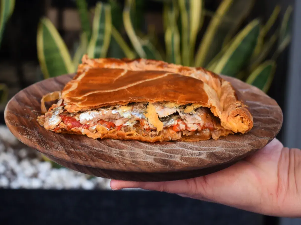 Una porción de empanada gallega, dorada y rellena de pescado y verduras, servida en plato de madera.