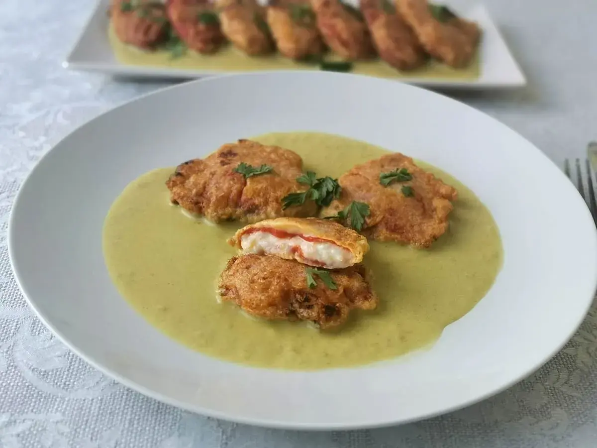Pimientos rellenos de bacalao rebozados, servidos en una cremosa salsa verde. Uno está cortado, mostrando el relleno blanco y rojo.