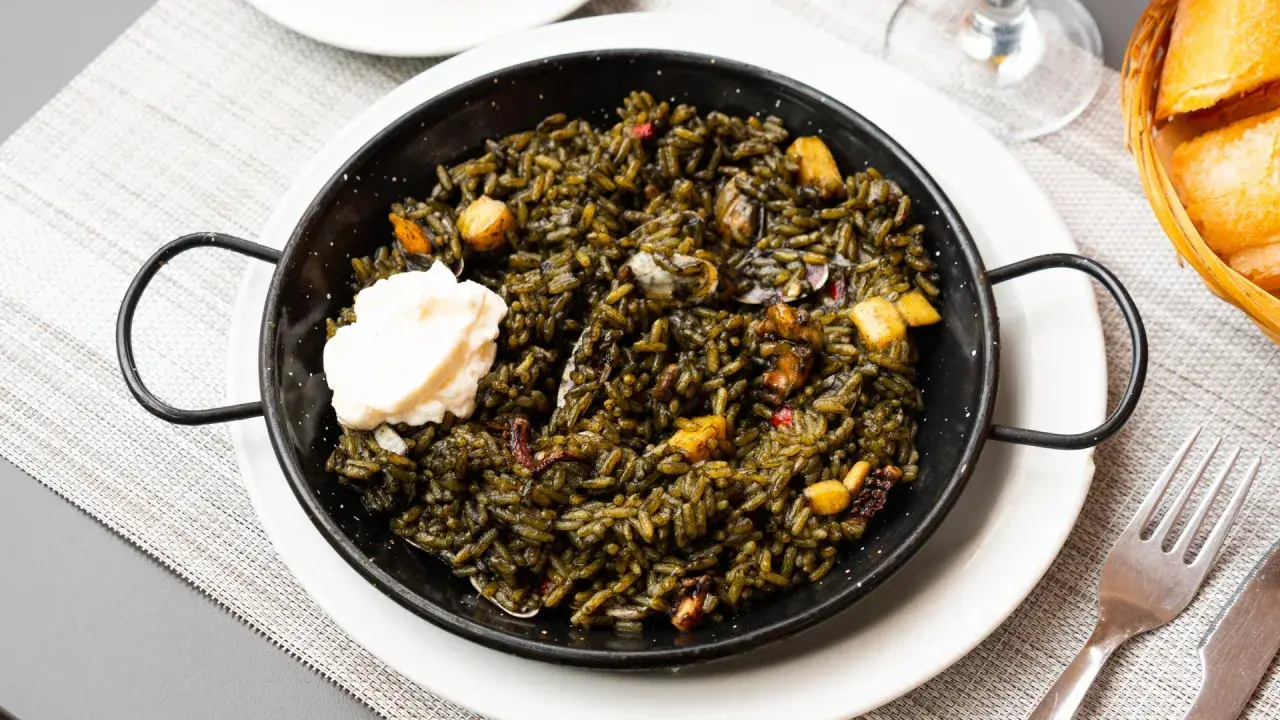 Arroz negro con sepia, servido en paellera con una cucharada de alioli. Se aprecian trozos de sepia y pimientos.